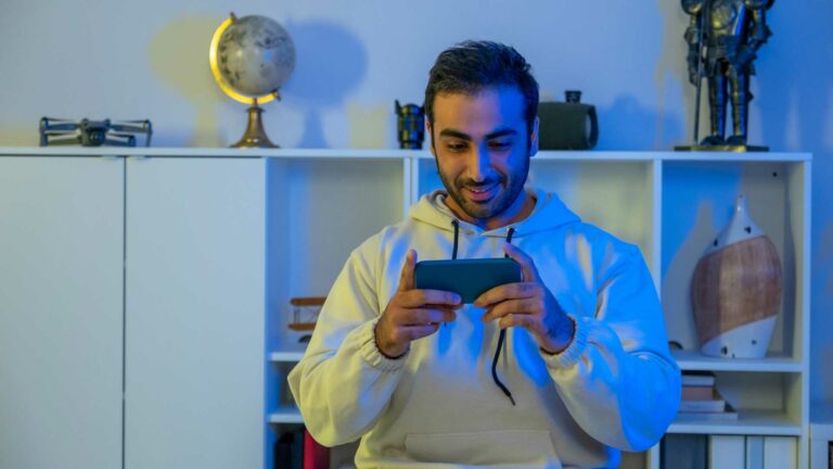 Homem sentado sorrindo e segurando o celular horizontalmente jogando um jogo.