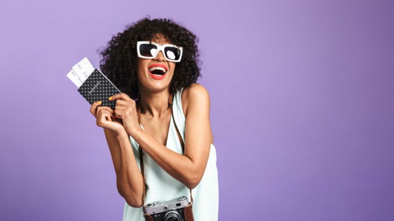 Mulher usando óculos escuros com câmera fotográfica no pescoço, segurando passaporte e passagens sorrindo